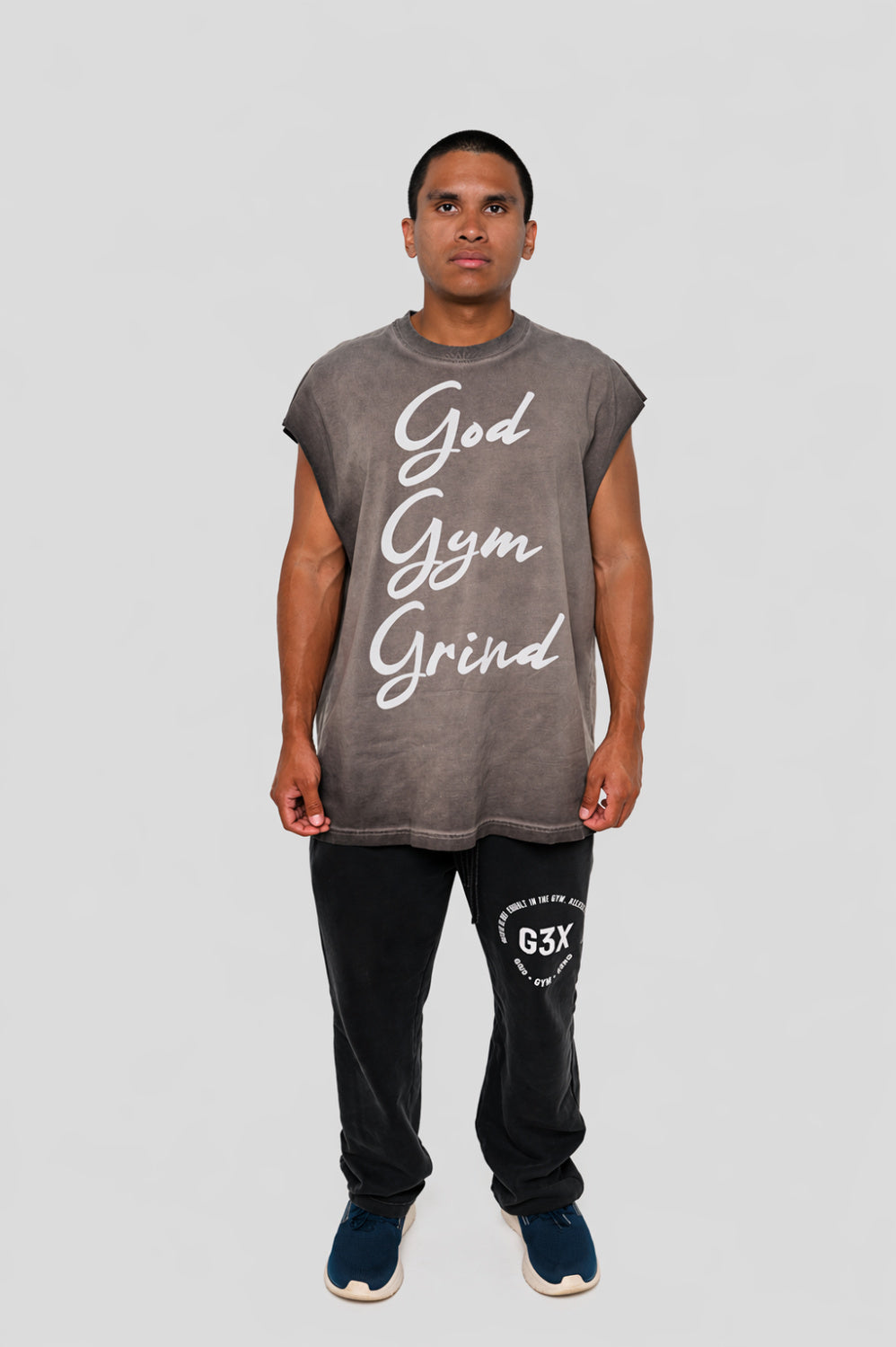 Man wearing a sleeveless gray shirt with 'God Gym Grind' text and black pants on a light gray background