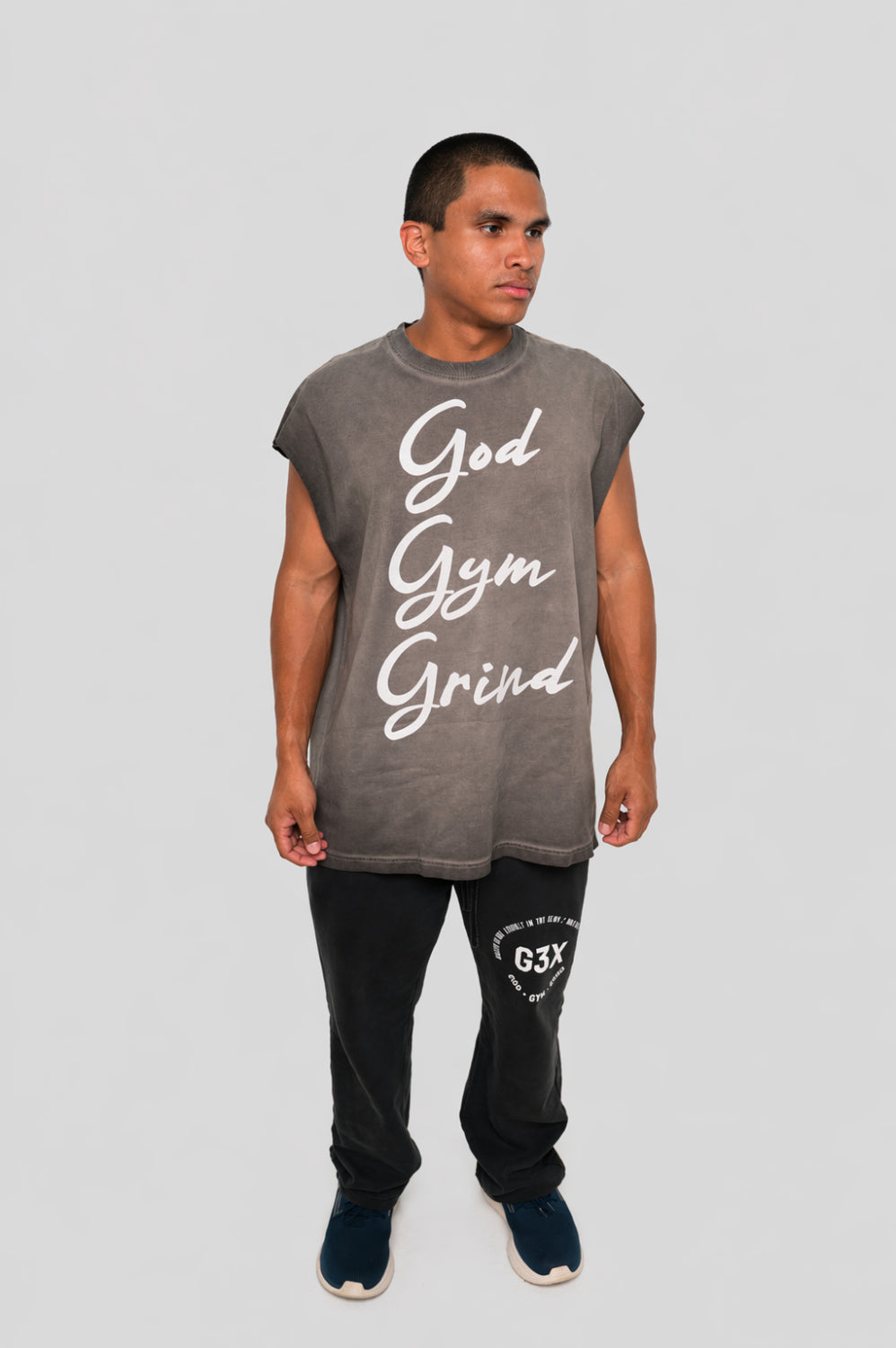 Man wearing a sleeveless gray shirt with 'God Gym Grind' text and black pants on a light gray background
